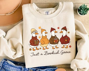 May include: Cream-colored sweatshirt with a graphic of four geese wearing hats and sweaters, each holding a book or a mug. The text "Just a Bookish Goose" is below the geese. Autumn leaves are scattered around the geese.