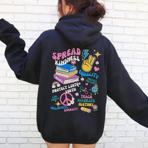 May include: Black hoodie with a colorful graphic print that says "Spread Kindness" and includes images of books, a peace sign, a lightning bolt, and a heart. The graphic also includes the phrases "Protect LGBTQ+ Youth", "Teach Accurate History", "Live Freely", "Protect the Earth", "Inclusivity", and "Diversity".