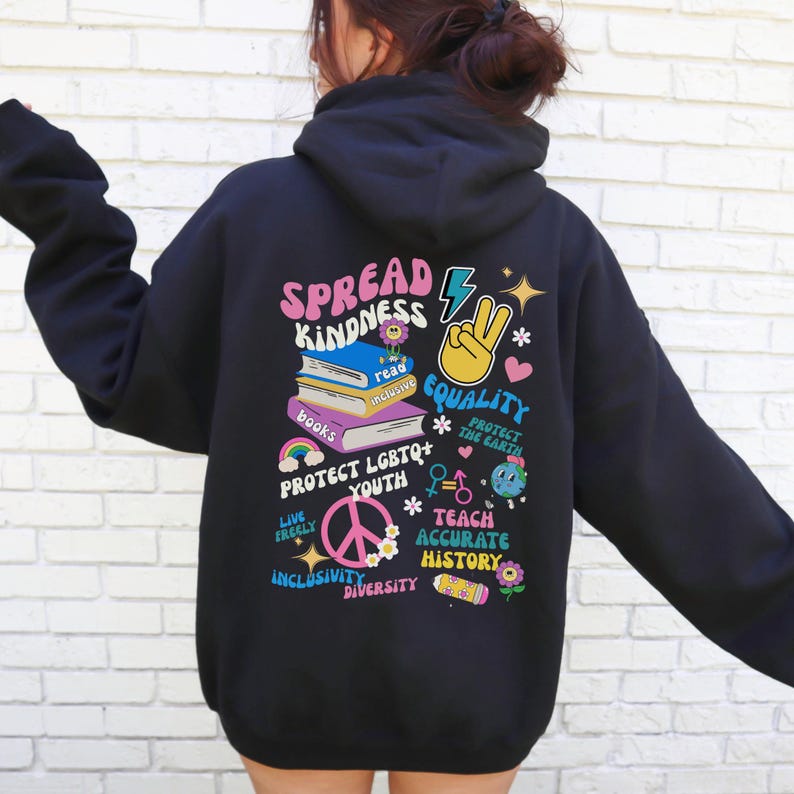 May include: Black hoodie with a colorful graphic print that says "Spread Kindness" and includes images of books, a peace sign, a lightning bolt, and a heart. The graphic also includes the phrases "Protect LGBTQ+ Youth", "Teach Accurate History", "Live Freely", "Protect the Earth", "Inclusivity", and "Diversity".
