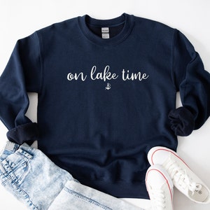 Puede incluir: Una sudadera azul marino con el texto "on lake time" en letras blancas y un peque&ntilde;o s&iacute;mbolo de ancla debajo del texto.
