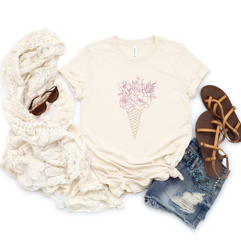 May include: A beige t-shirt with a pink floral design in the shape of an ice cream cone. The shirt is laid flat on a white background with a pair of brown sandals and a white crocheted scarf.