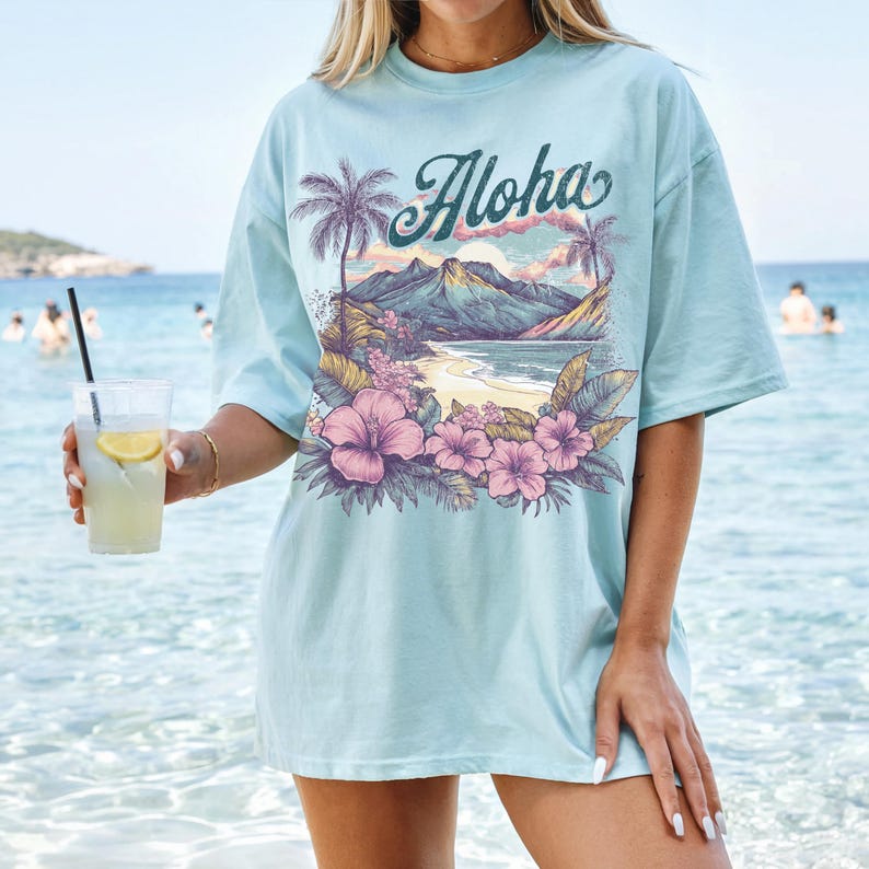 以下が含まれることがあります： 「アロハ」の文字が書かれた水色のTシャツ。ヤシの木、山、ピンクの花が描かれたトロピカルな風景が特徴です。ビーチで着用されています。