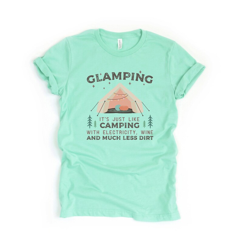 May include: A light green t-shirt with the text "GLAMPING" in black letters. The text "IT'S JUST LIKE CAMPING WITH ELECTRICITY, WINE AND MUCH LESS DIRT" is printed below the word "GLAMPING". A cartoon illustration of a tent with string lights and a sleeping bag inside is shown above the text.