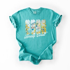 Pu&ograve; includere: Una t-shirt blu turchese con una stampa grafica di una citt&agrave; colorata con alberi di limone e il testo "Amalfi Coast".