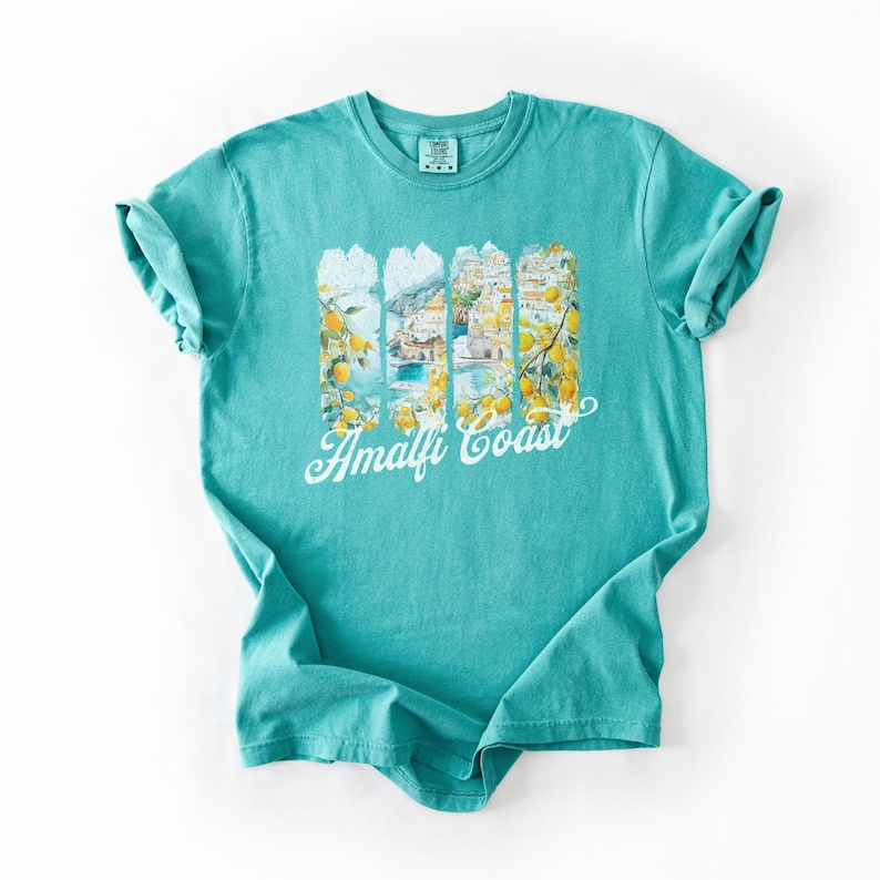 Pu&ograve; includere: Una t-shirt blu turchese con una stampa grafica di una citt&agrave; colorata con alberi di limone e il testo "Amalfi Coast".