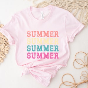 May include: A light pink t-shirt with the word "SUMMER" printed four times in different colors: coral, yellow, teal, and fuchsia. The shirt is knotted at the bottom and has rolled-up sleeves.