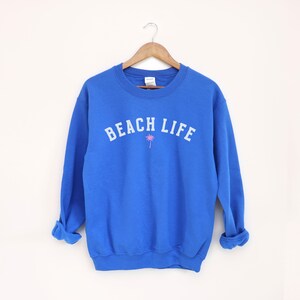 Pode incluir: Uma camisola azul real com o texto "BEACH LIFE" em branco e um gr&aacute;fico de palmeira rosa.
