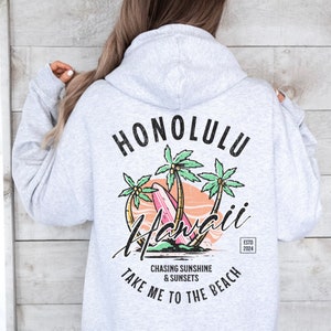 May include: Gray hooded sweatshirt with a graphic print of palm trees, a surfboard, and the text "Honolulu Hawaii" in black. The text "Chasing Sunshine & Sunsets Take Me To The Beach" is printed below the graphic. The text "Estd 2024" is printed in a small rectangle in the bottom right corner of the graphic.