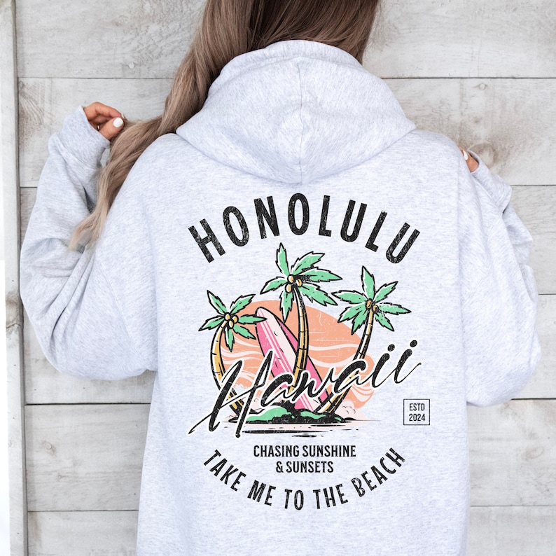 May include: Gray hooded sweatshirt with a graphic print of palm trees, a surfboard, and the text "Honolulu Hawaii" in black. The text "Chasing Sunshine & Sunsets Take Me To The Beach" is printed below the graphic. The text "Estd 2024" is printed in a small rectangle in the bottom right corner of the graphic.