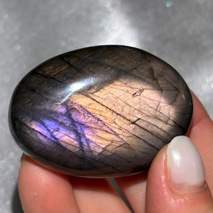 May include: A smooth, oval labradorite stone. The stone has a dark grey base with iridescent flashes of purple, gold, and blue. The surface is polished and reflective, displaying natural patterns and colour variations.