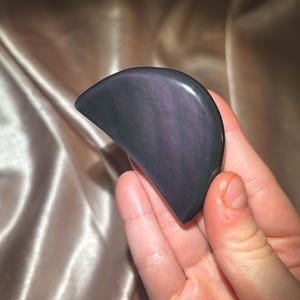 Może przedstawiać: Narzędzie do gua sha z obsydianu w kształcie p&oacute;łksiężyca z tęczowymi odcieniami fioletu i czerni. Narzędzie jest trzymane w dłoni.