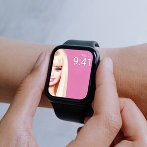 Puede incluir: Un reloj inteligente negro con una esfera rosa que muestra la hora 9:41 y la fecha MAR 12. La esfera tambi&eacute;n presenta una imagen de una mu&ntilde;eca Barbie.