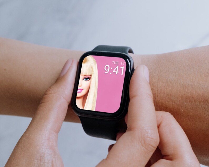 Puede incluir: Un reloj inteligente negro con una esfera rosa que muestra la hora 9:41 y la fecha MAR 12. La esfera tambi&eacute;n presenta una imagen de una mu&ntilde;eca Barbie.