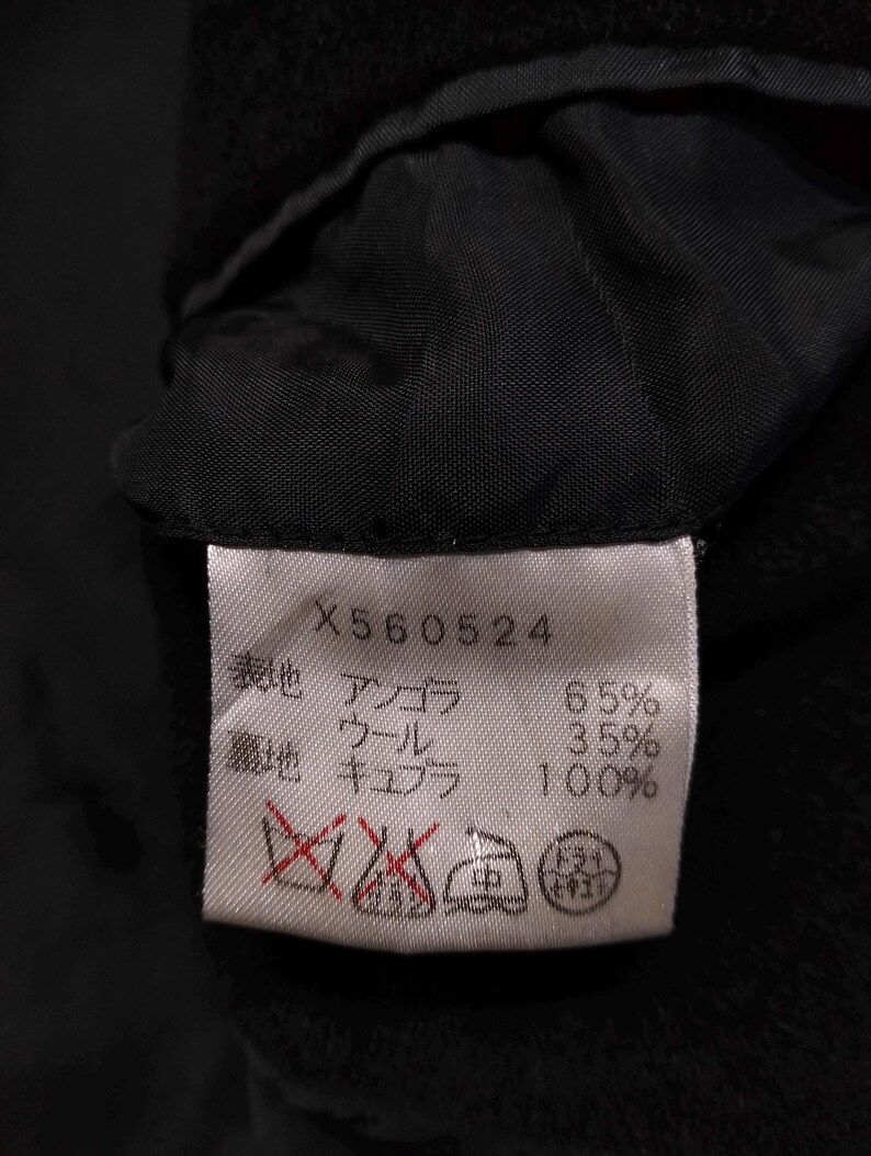 May include: A white rectangular garment care label with black text and symbols sewn into a black fabric lining. The label displays the code X560524, material composition of 65% angora and 35% wool for the outer shell, 100% cupro for the lining, and four care icons.