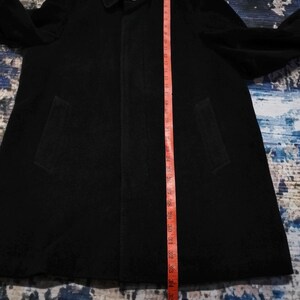 May include: A black coat with a collar, long sleeves, and two side pockets. A red measuring tape is placed vertically along the front, showing a length of approximately 38 inches from the collar down to the bottom hem of the garment.