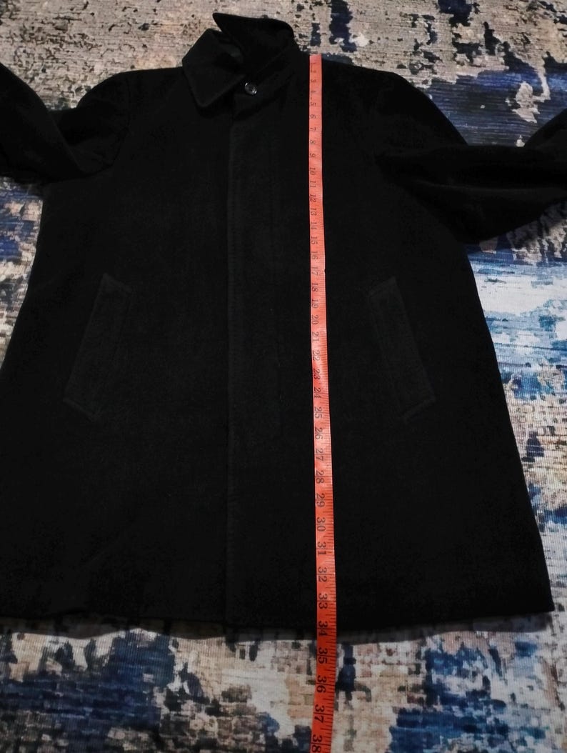 May include: A black coat with a collar, long sleeves, and two side pockets. A red measuring tape is placed vertically along the front, showing a length of approximately 38 inches from the collar down to the bottom hem of the garment.