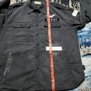 Puede incluir: Una camisa gris oscuro con botones, dos bolsillos en el pecho y dos bolsillos inferiores. La camisa presenta parches con texto y un emblema alado. El texto incluye "U.S.A.A.F." y "CONTRACT W 535 AC 23619". Una cinta m&eacute;trica se coloca verticalmente.