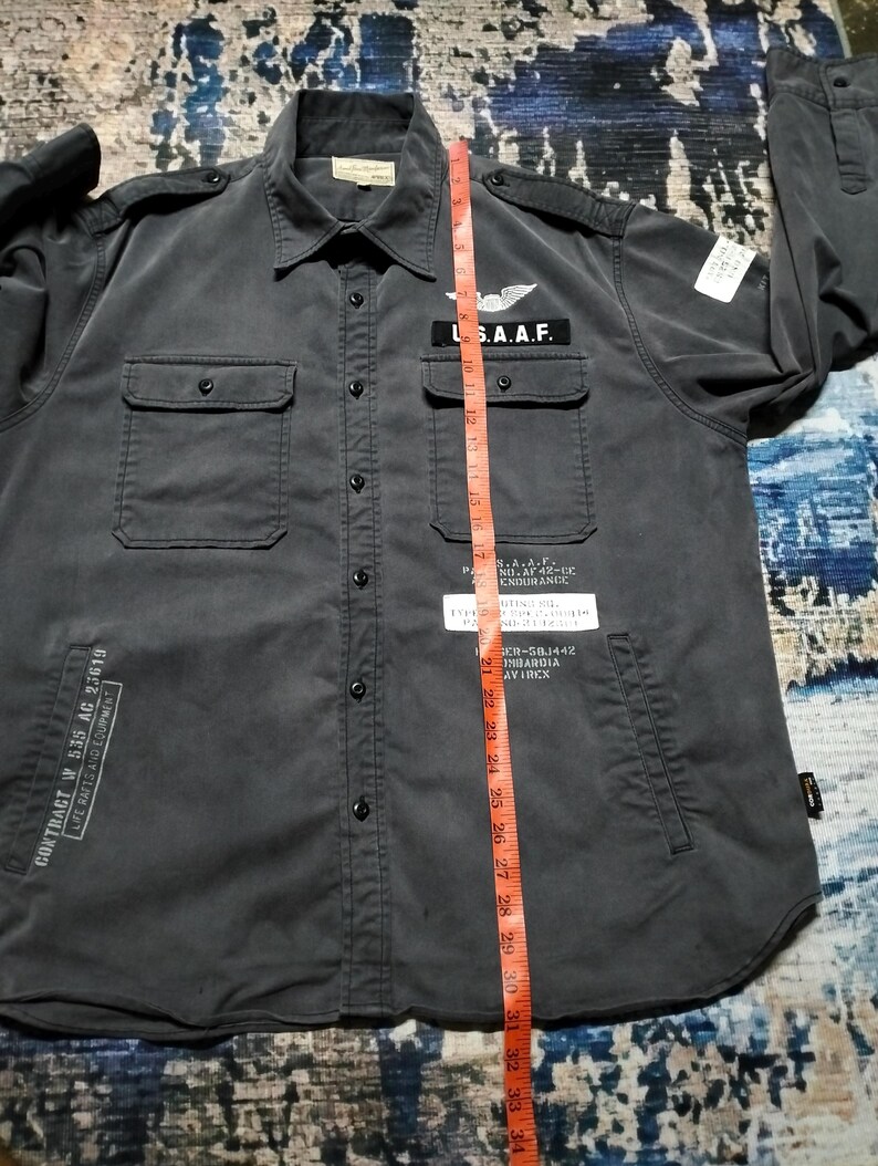 Puede incluir: Una camisa gris oscuro con botones, dos bolsillos en el pecho y dos bolsillos inferiores. La camisa presenta parches con texto y un emblema alado. El texto incluye "U.S.A.A.F." y "CONTRACT W 535 AC 23619". Una cinta m&eacute;trica se coloca verticalmente.