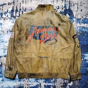 May include: A tan leather jacket featuring a large graphic on the back with a map of the United States and the red text American Born. The jacket includes a small American flag patch on the left sleeve, an Adidas logo patch, and a circular emblem.