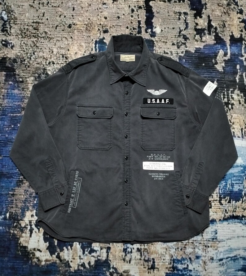 Puede incluir: Camisa abotonada gris oscuro con dos bolsillos en el pecho y parches. La camisa presenta el texto "U.S.A.A.F." y otro texto de estilo militar. La camisa tiene mangas largas y cuello.