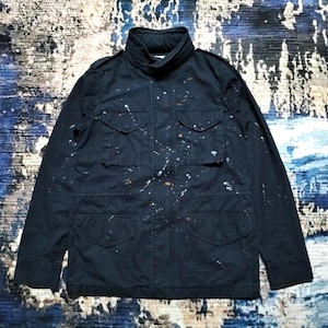 May include: A dark navy blue field jacket featuring a high collar, long sleeves, and four front pockets with buttoned flaps. The jacket is decorated with a scattered white and light brown paint splatter pattern across the front and sleeves.