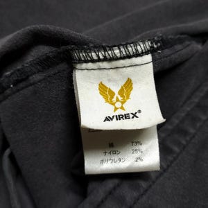 Puede incluir: Primer plano de una etiqueta de prenda gris oscuro con la marca AVIREX y un logotipo de &aacute;guila dorada. La etiqueta tambi&eacute;n enumera la composici&oacute;n de la tela: 73% algod&oacute;n, 25% nailon y 2% poliuretano.