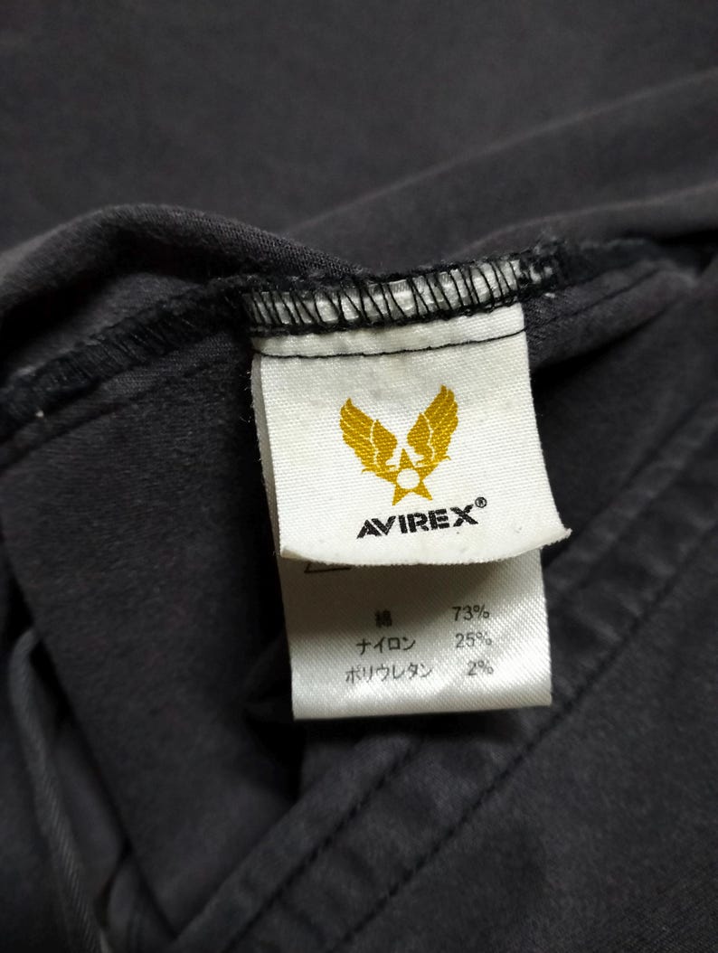 Puede incluir: Primer plano de una etiqueta de prenda gris oscuro con la marca AVIREX y un logotipo de &aacute;guila dorada. La etiqueta tambi&eacute;n enumera la composici&oacute;n de la tela: 73% algod&oacute;n, 25% nailon y 2% poliuretano.