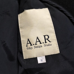 May include: A black garment interior featuring a rectangular cream-colored fabric label. The label displays the text A.A.R and Yohji Design Studio in black lettering. A smaller white tag below the main label indicates the size as M.