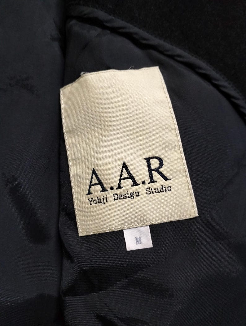 May include: A black garment interior featuring a rectangular cream-colored fabric label. The label displays the text A.A.R and Yohji Design Studio in black lettering. A smaller white tag below the main label indicates the size as M.