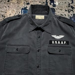 Puede incluir: Camisa gris oscuro con botones, dos bolsillos en el pecho y charreteras. Presenta un parche que dice "U.S.A.A.F." encima del bolsillo derecho y un emblema de ala. La camisa tiene cuello y una etiqueta en el cuello.