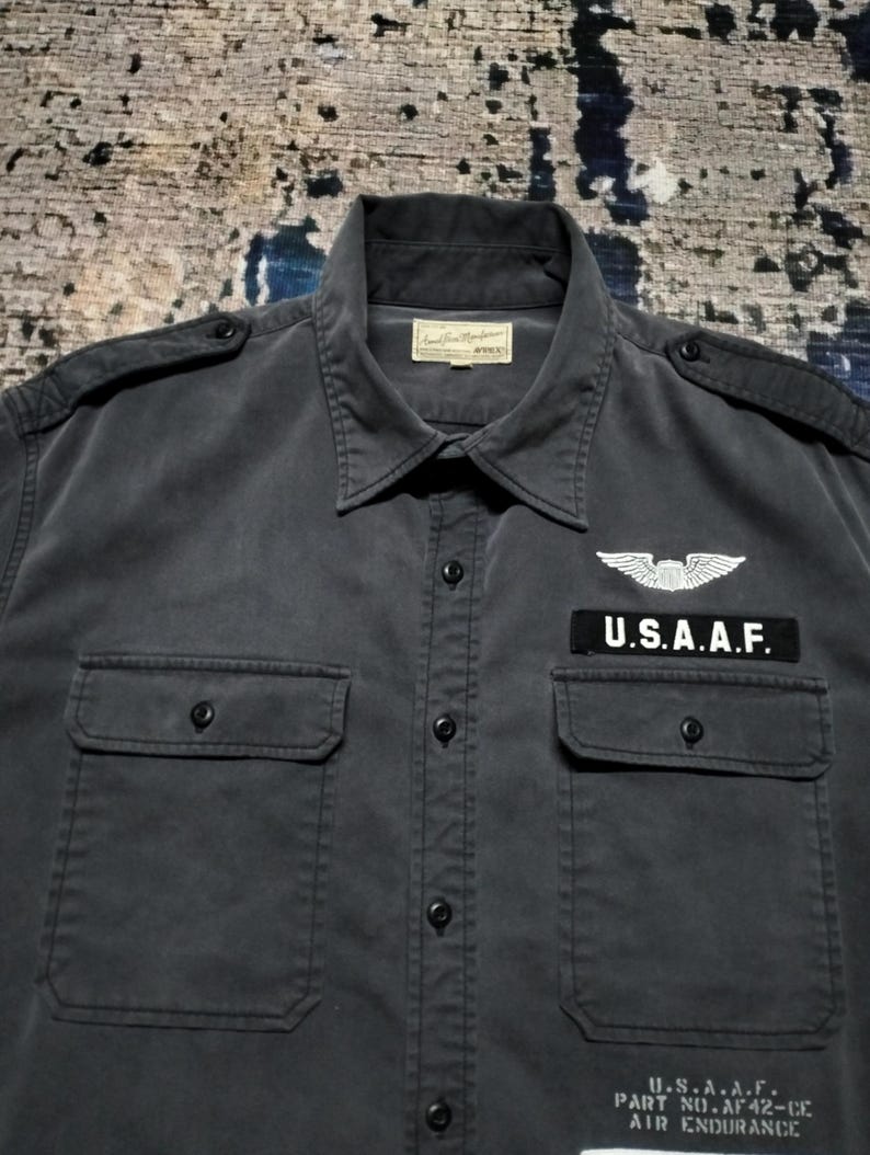Puede incluir: Camisa gris oscuro con botones, dos bolsillos en el pecho y charreteras. Presenta un parche que dice "U.S.A.A.F." encima del bolsillo derecho y un emblema de ala. La camisa tiene cuello y una etiqueta en el cuello.