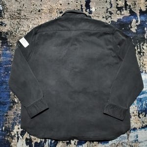 Puede incluir: Una camisa gris oscuro de manga larga con cuello abotonado y pu&ntilde;os. La camisa tiene una peque&ntilde;a etiqueta blanca en el hombro izquierdo. La tela parece ser un algod&oacute;n o mezcla resistente, con un aspecto ligeramente desgastado.