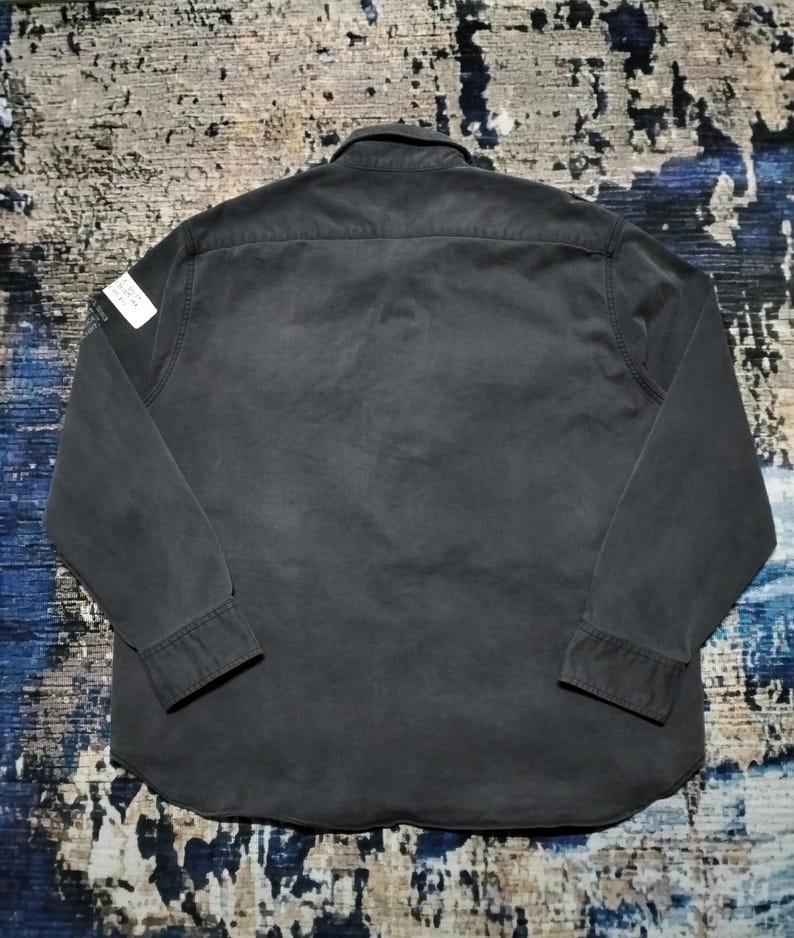 Puede incluir: Una camisa gris oscuro de manga larga con cuello abotonado y pu&ntilde;os. La camisa tiene una peque&ntilde;a etiqueta blanca en el hombro izquierdo. La tela parece ser un algod&oacute;n o mezcla resistente, con un aspecto ligeramente desgastado.
