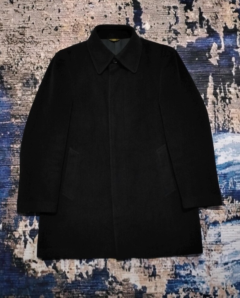 May include: A solid black coat featuring a structured collar, long sleeves, and a concealed button placket. The garment has a clean, minimalist silhouette with two side pockets and a smooth, dark fabric finish, presenting a classic design.