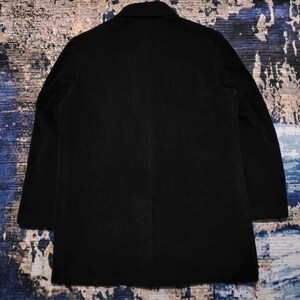 May include: A solid black blazer featuring a structured collar, long sleeves, and a straight hemline. The garment is displayed from the back, showcasing a clean, minimalist design with a single vertical seam running down the center of the back panel.