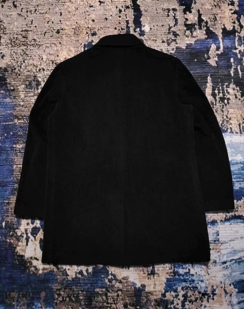 May include: A solid black blazer featuring a structured collar, long sleeves, and a straight hemline. The garment is displayed from the back, showcasing a clean, minimalist design with a single vertical seam running down the center of the back panel.