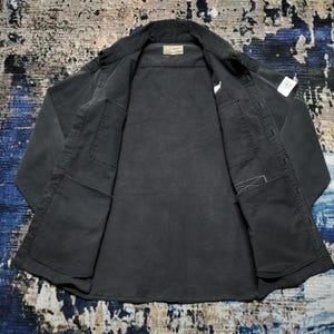 Puede incluir: Una camisa gris oscuro con botones, dos bolsillos delanteros y un cuello. La camisa est&aacute; abierta, revelando el forro interior y una peque&ntilde;a etiqueta cerca del cuello. La tela parece ser un material resistente, posiblemente de estilo workwear.