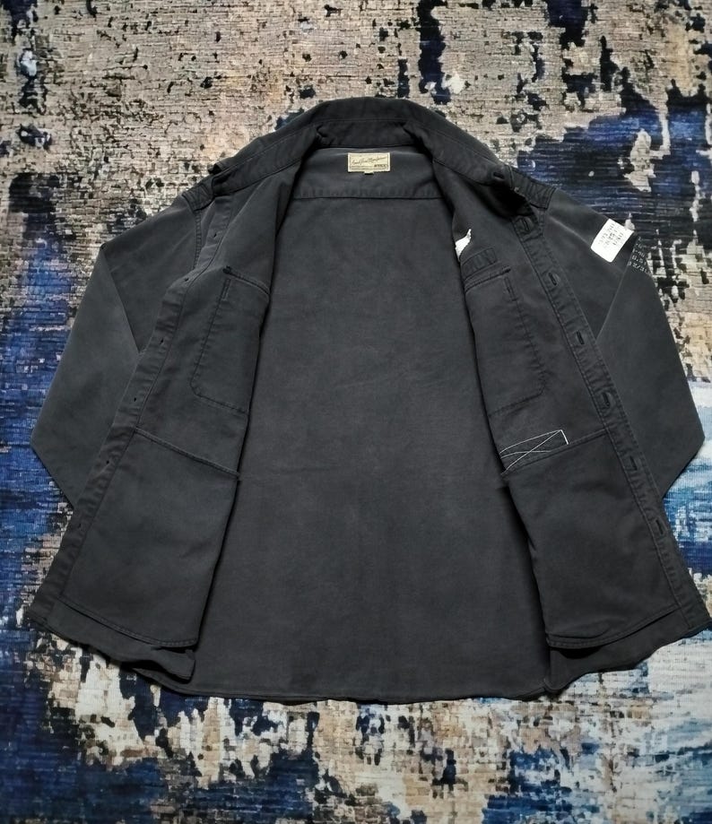 Puede incluir: Una camisa gris oscuro con botones, dos bolsillos delanteros y un cuello. La camisa est&aacute; abierta, revelando el forro interior y una peque&ntilde;a etiqueta cerca del cuello. La tela parece ser un material resistente, posiblemente de estilo workwear.