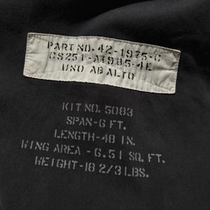 Puede incluir: Primer plano de una tela oscura con texto impreso. El texto incluye un n&uacute;mero de pieza, n&uacute;mero de kit, envergadura, longitud (122 cm), superficie alar y peso (8,4 kg). La tela es de color gris oscuro o negro.