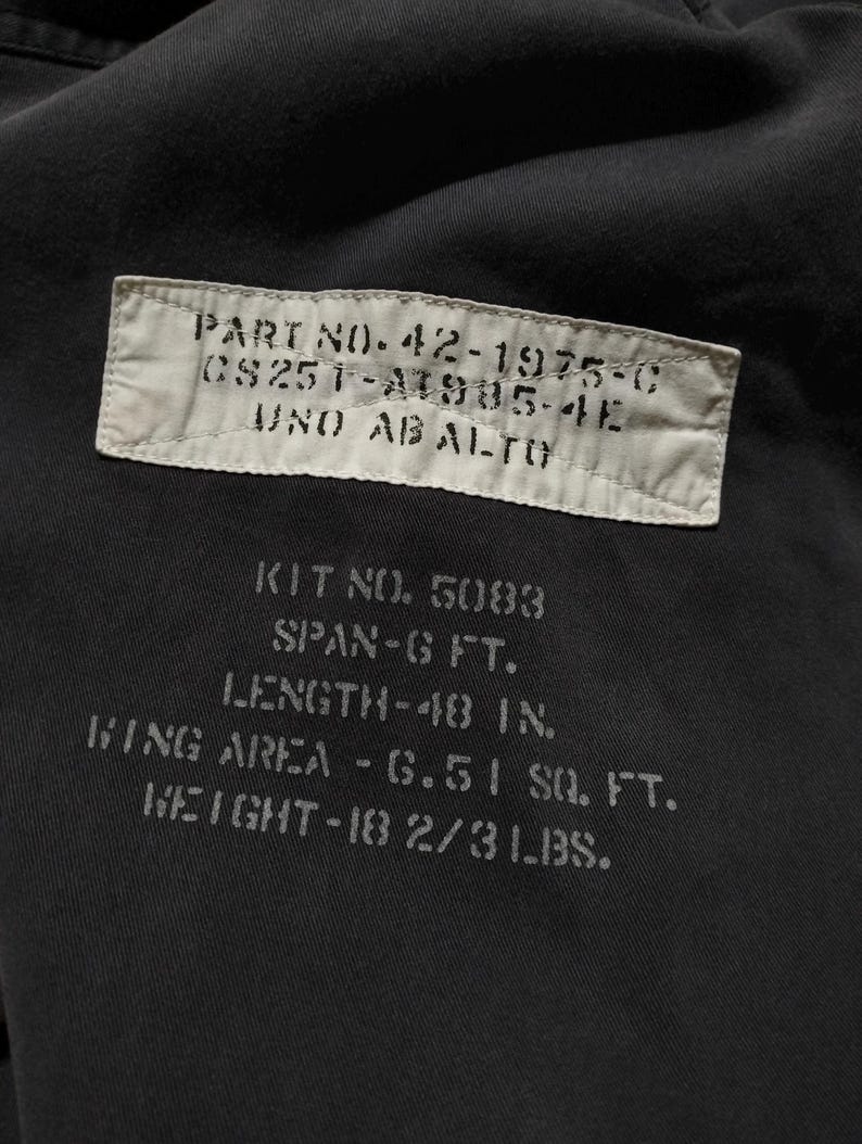 Puede incluir: Primer plano de una tela oscura con texto impreso. El texto incluye un n&uacute;mero de pieza, n&uacute;mero de kit, envergadura, longitud (122 cm), superficie alar y peso (8,4 kg). La tela es de color gris oscuro o negro.