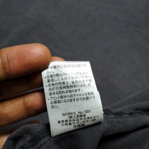 Puede incluir: Primer plano de una tela gris oscuro con una etiqueta blanca adjunta. La etiqueta tiene texto japon&eacute;s y las palabras "WORKS No. 001". La etiqueta tambi&eacute;n incluye instrucciones de lavado.