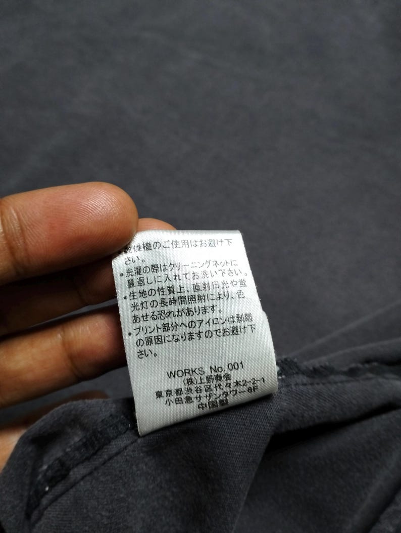 Puede incluir: Primer plano de una tela gris oscuro con una etiqueta blanca adjunta. La etiqueta tiene texto japon&eacute;s y las palabras "WORKS No. 001". La etiqueta tambi&eacute;n incluye instrucciones de lavado.