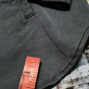 Puede incluir: Primer plano de una tela gris oscuro con costuras visibles y un peque&ntilde;o detalle de bolsillo. Una cinta m&eacute;trica de fibra de vidrio roja y blanca est&aacute; colocada sobre la tela.