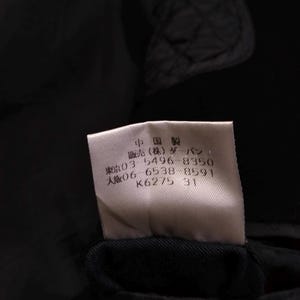 May include: A white rectangular garment care label sewn onto a black quilted fabric. The label features black Japanese text including "中国製", "販売(株)ダーバン", and phone numbers "03 5496 8350" and "06 6538 8591", along with the alphanumeric code "K6275 31".