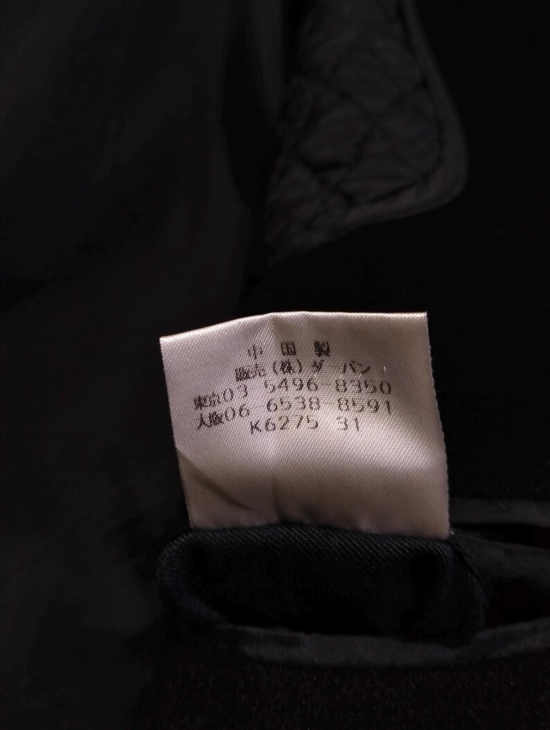 May include: A white rectangular garment care label sewn onto a black quilted fabric. The label features black Japanese text including "中国製", "販売(株)ダーバン", and phone numbers "03 5496 8350" and "06 6538 8591", along with the alphanumeric code "K6275 31".