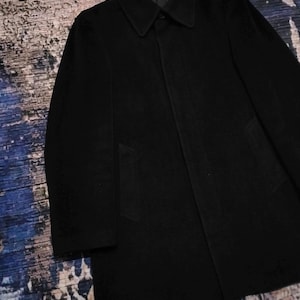 May include: A solid black coat featuring a structured collar, long sleeves, and a concealed button placket. The garment has a smooth, matte texture and a straight hemline, with two side pockets integrated into the front panel of the coat.