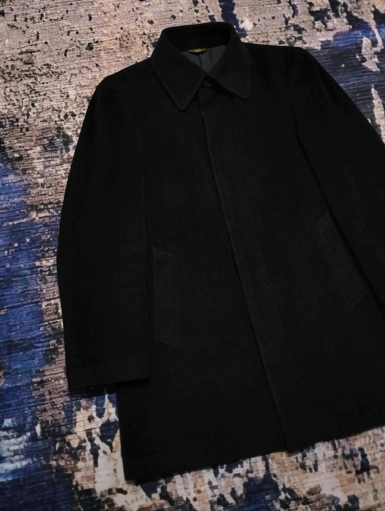 May include: A solid black coat featuring a structured collar, long sleeves, and a concealed button placket. The garment has a smooth, matte texture and a straight hemline, with two side pockets integrated into the front panel of the coat.