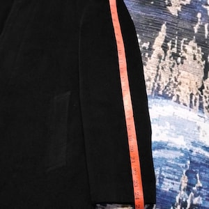 May include: A black coat with a long sleeve, featuring a visible button at the collar and a side pocket. A pink measuring tape is placed along the sleeve, indicating a length of approximately 27 inches from the shoulder seam to the cuff edge.