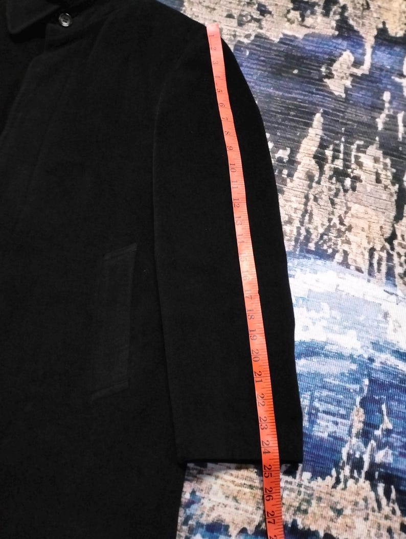 May include: A black coat with a long sleeve, featuring a visible button at the collar and a side pocket. A pink measuring tape is placed along the sleeve, indicating a length of approximately 27 inches from the shoulder seam to the cuff edge.