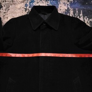 May include: A solid black coat with a pointed collar and long sleeves. A red measuring tape is placed horizontally across the chest, showing a width measurement of approximately 26 inches from one side to the other across the front of the garment.